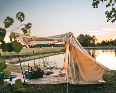 A Nordisk glamping tent with a canopy and fairy lights by a lake at sunset.