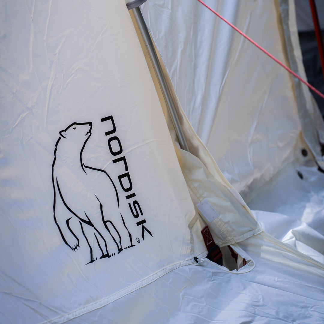 Close-up of a white tent fabric with a black outlined polar bear logo and the word NORDISK printed vertically, along with part of the tent pole and a red guyline.