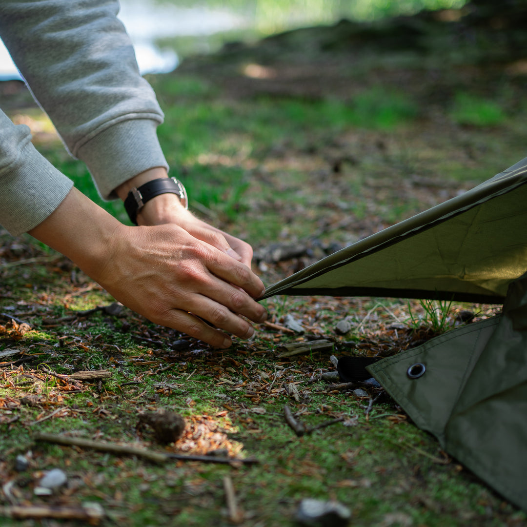A close-up lifestyle shot of a person's hands securing an olive green tent peg into the ground outdoors on a forest floor.