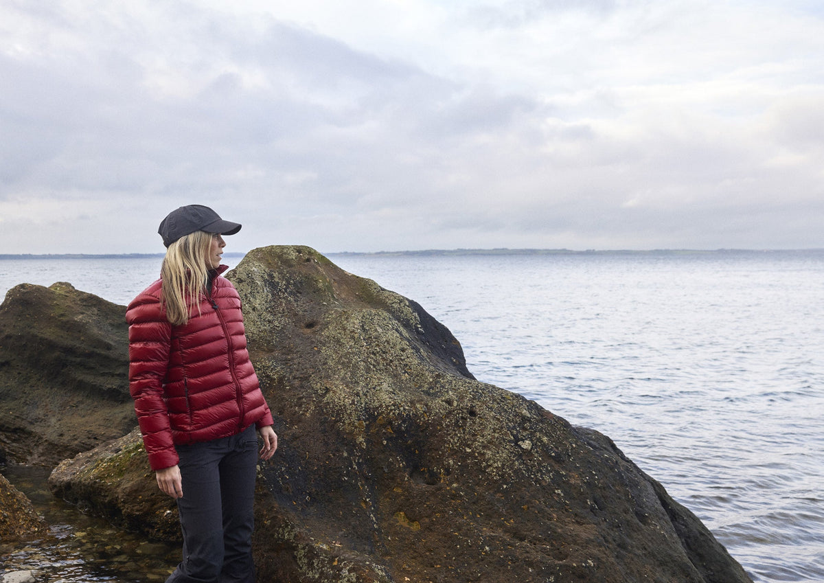 Woman wearing the Companyon Thyra down jacket in Tibetan Red on the rocks by the sea