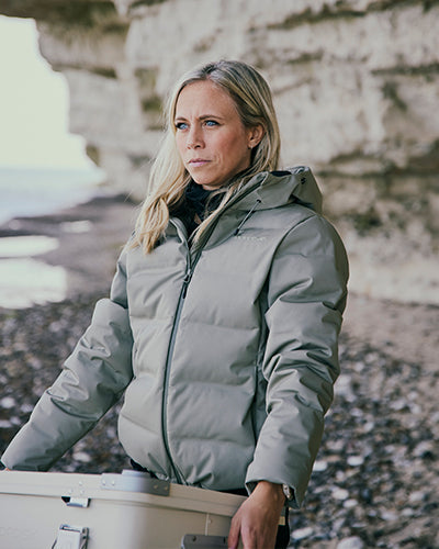 Woman wearing a grey Nordisk down jacket on a rocky beach by the ocean.