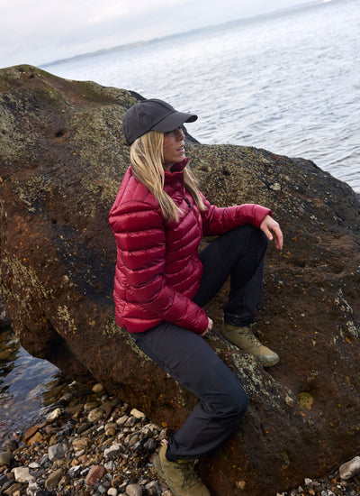 Woman wearing the Companyon Thyra down jacket in Tibetan Red on a rock by the sea