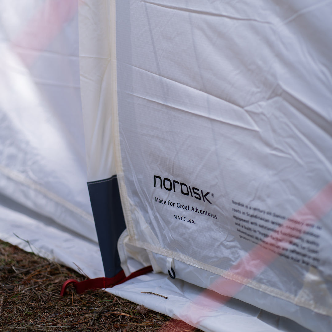 Close-up of a white Nordisk tent fabric with printed brand text, shown outdoors with ground visible at the bottom and a red guy line attached.