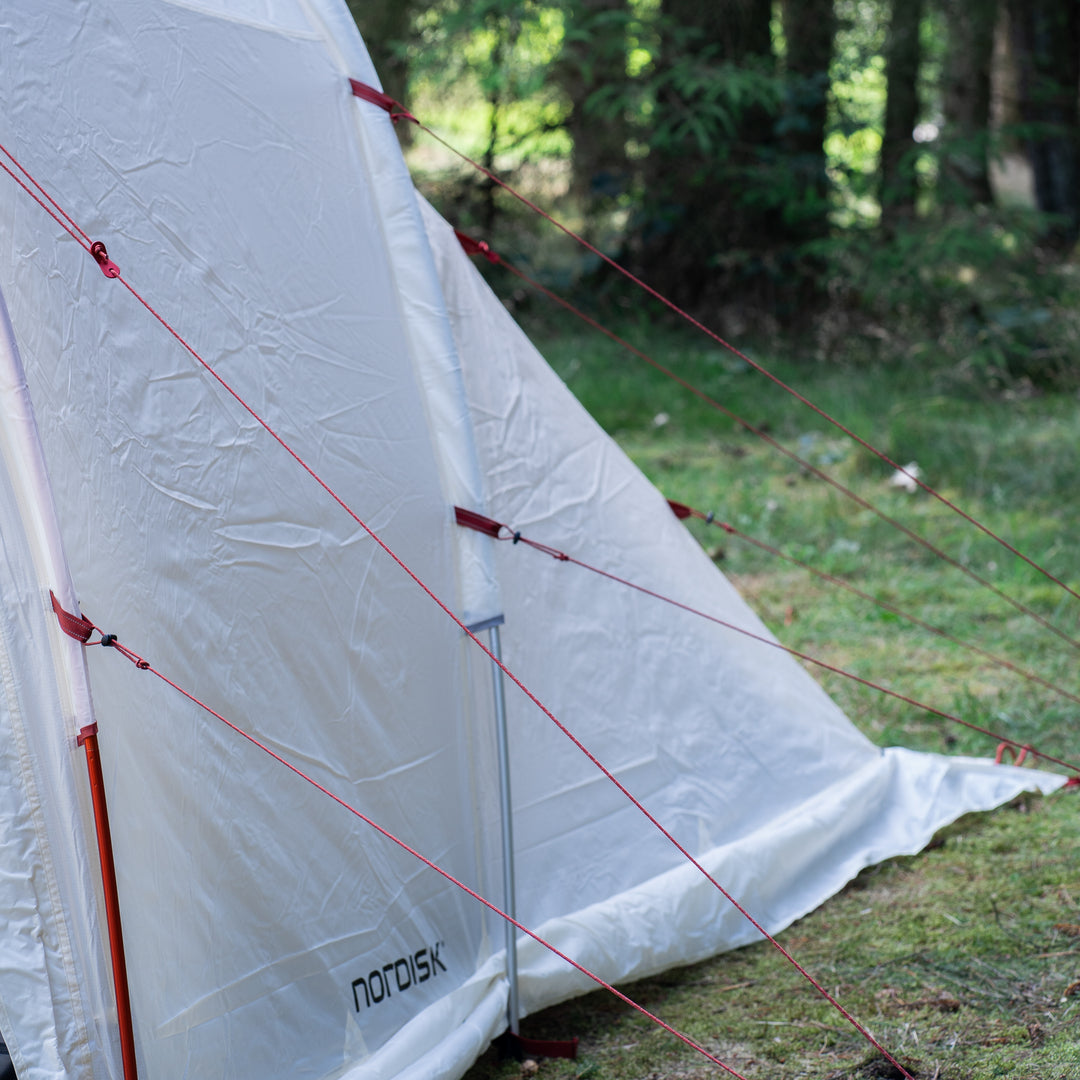 A white Nordisk camping tent pitched outdoors on grass with trees in the background, showing red guy lines and tent poles.