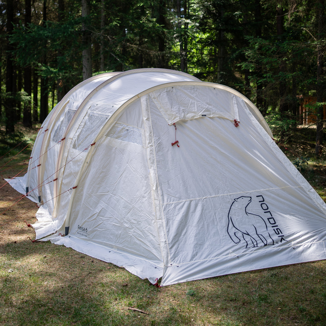 A white Nordisk dome tent pitched outdoors in a wooded clearing with trees and grass visible around it.