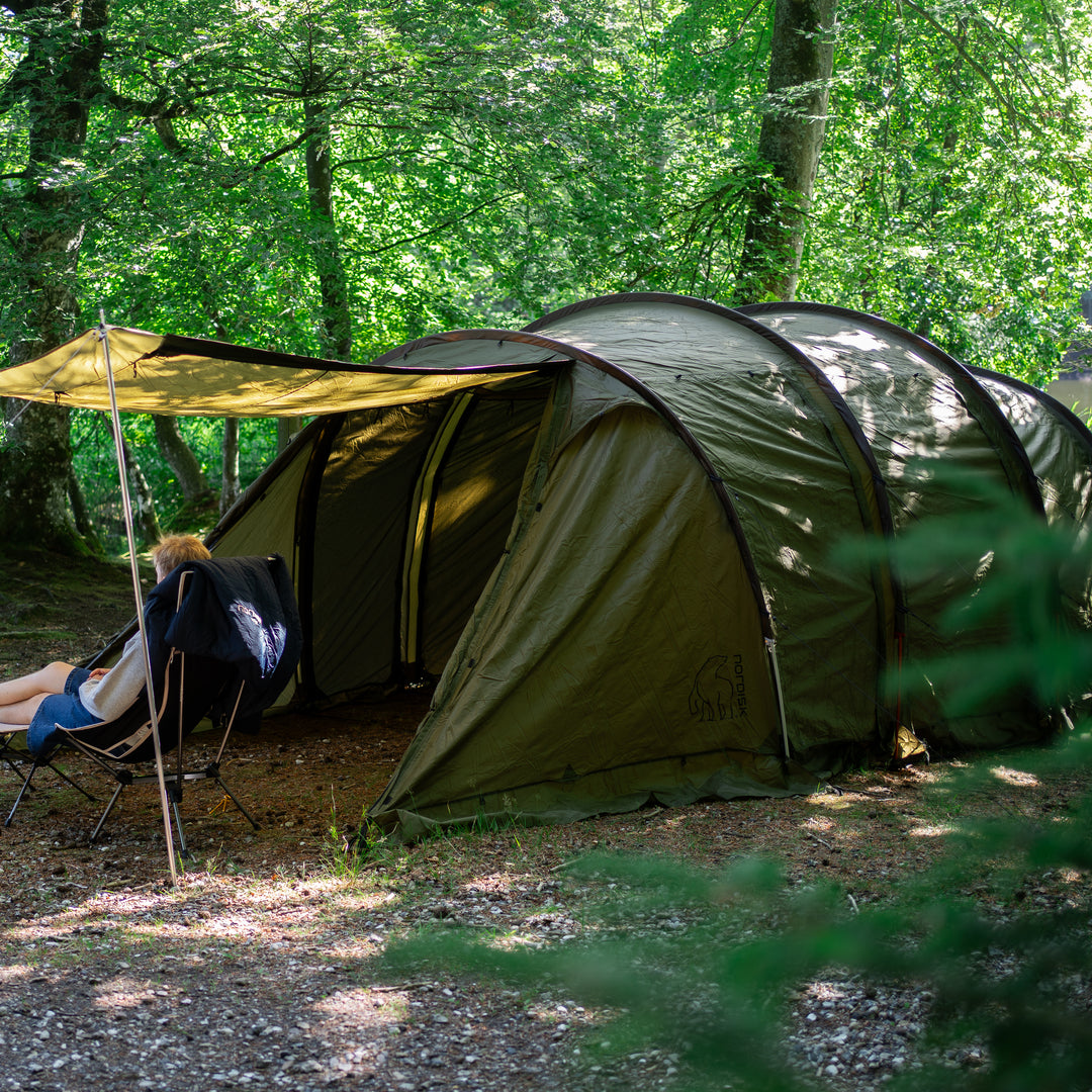 An olive green dome camping tent pitched in a wooded forest clearing, with a person sitting in a folding chair under a yellow tarp shelter.