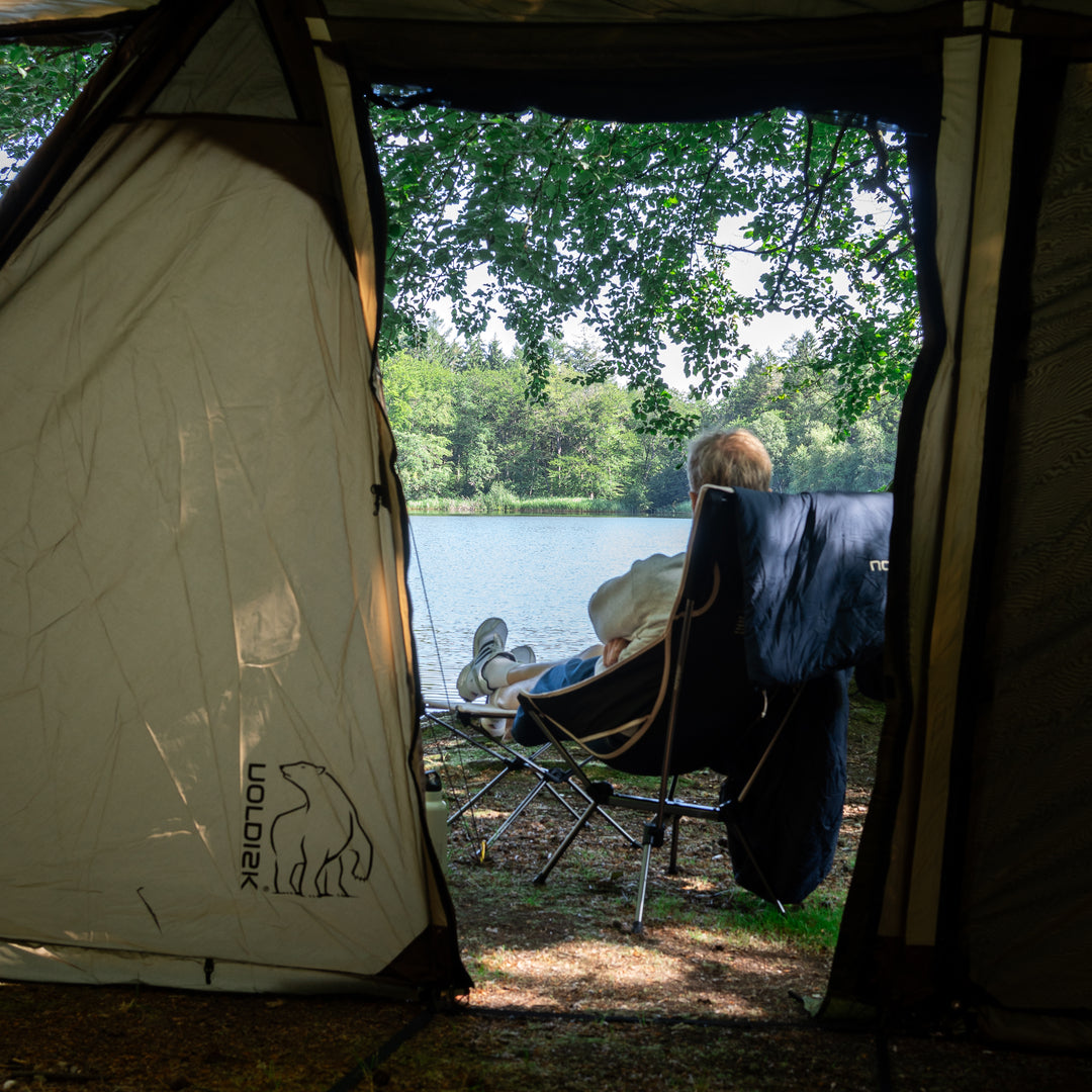 A person relaxes in a black camping chair inside a tent, with a lake and wooded shoreline visible outside. The tent opening is framed prominently, and the brand name Nordisk with a polar bear logo is printed on the left side of the tent.