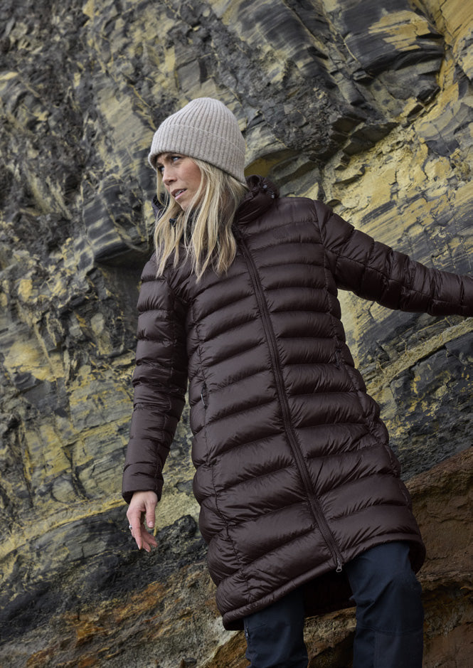 Woman wearing the Companyon Dagmar down coat in Black Coffee by a rock face